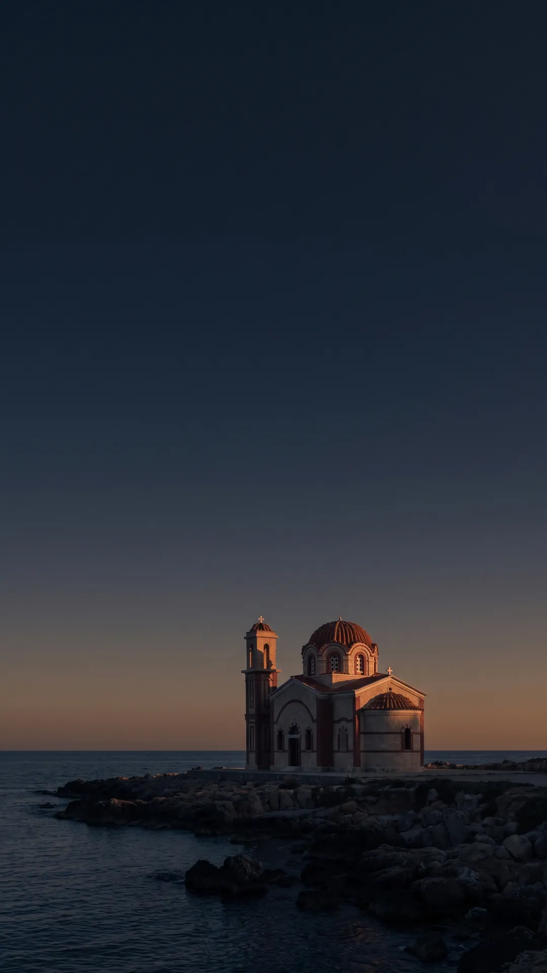 A small Cypriot chapel with a red-tiled dome and bell tower on a rocky shoreline at dusk