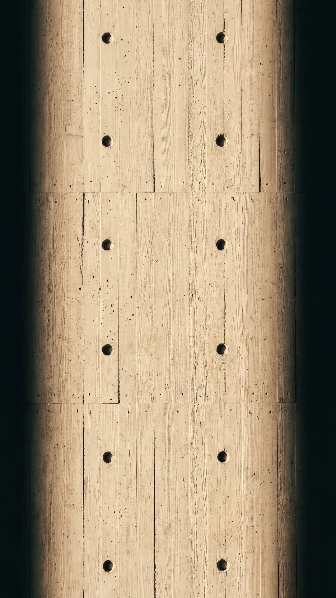 Fair-faced concrete with a grid of form-tie holes under raking warm light
