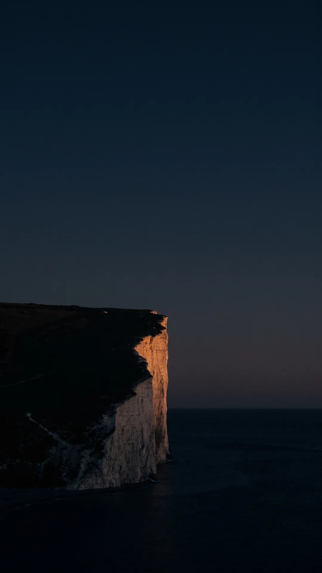 A dark sea cliff at dusk, pale limestone catching the last warm light